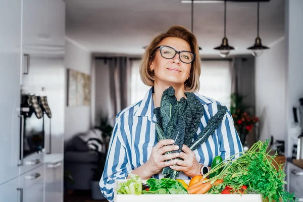 Smiling woman holding fresh kale from her healthy food delivery. Wooden box with vegetables. Support Local farmer food. Start of a healthy life, weight loss concept. Online home food delivery. — Stock Photo, Image