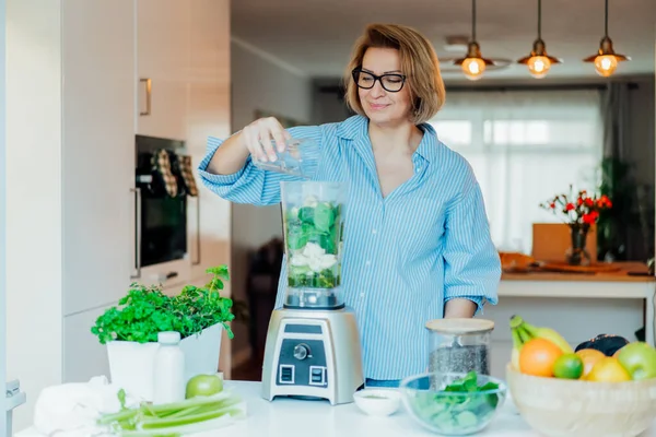Middle aged woman with blender and green vegetables making detox shake or green smoothie at home. Healthy dieting, eating, cooking. Natural anti aging, weight loss program. Vegan, vegetarian diet — 스톡 사진