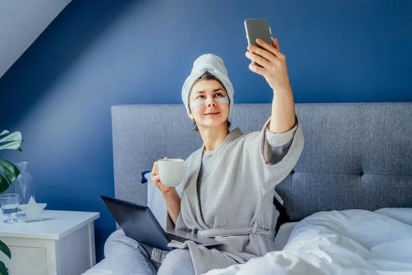 Woman in pajama and bathrobe with eyes patches and towel on her head drinking morning coffee and making selfie by phone on her bed. Morning habits and home beauty routine. Content creating