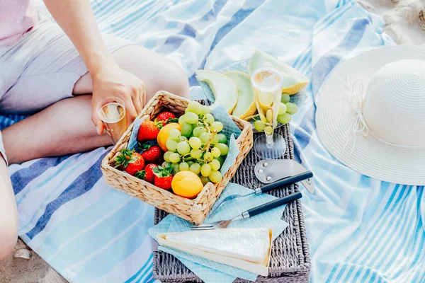 Sommer-Picknick am Strand bei Sonnenuntergang. Junger Mann sitzt auf einer Decke und picknickt am Wochenende draußen am Meer mit frischem Obst der Saison, Käse und Sektgläsern. Romantisches Date für zwei. Ansicht von oben. — Stockfoto