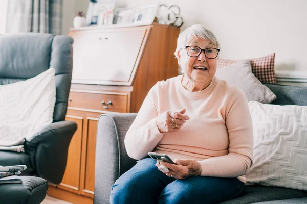 Retrato de mujer mayor riendo feliz usando el teléfono mientras está sentada en el sofá en casa. Señora mayor madura está emocionado de recibir buenas noticias a través de teléfono inteligente. Mayores y tecnología. Enfoque selectivo. —  Fotos de Stock