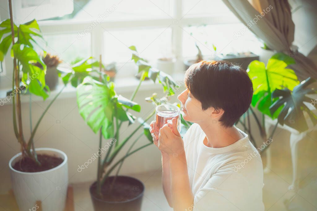 Woman with closed eyes drinking tea in a room with green house plants, enjoying its taste and aroma in a sunset or sunrise light. Relaxing and self-care, personal fulfillment. Selective focus.