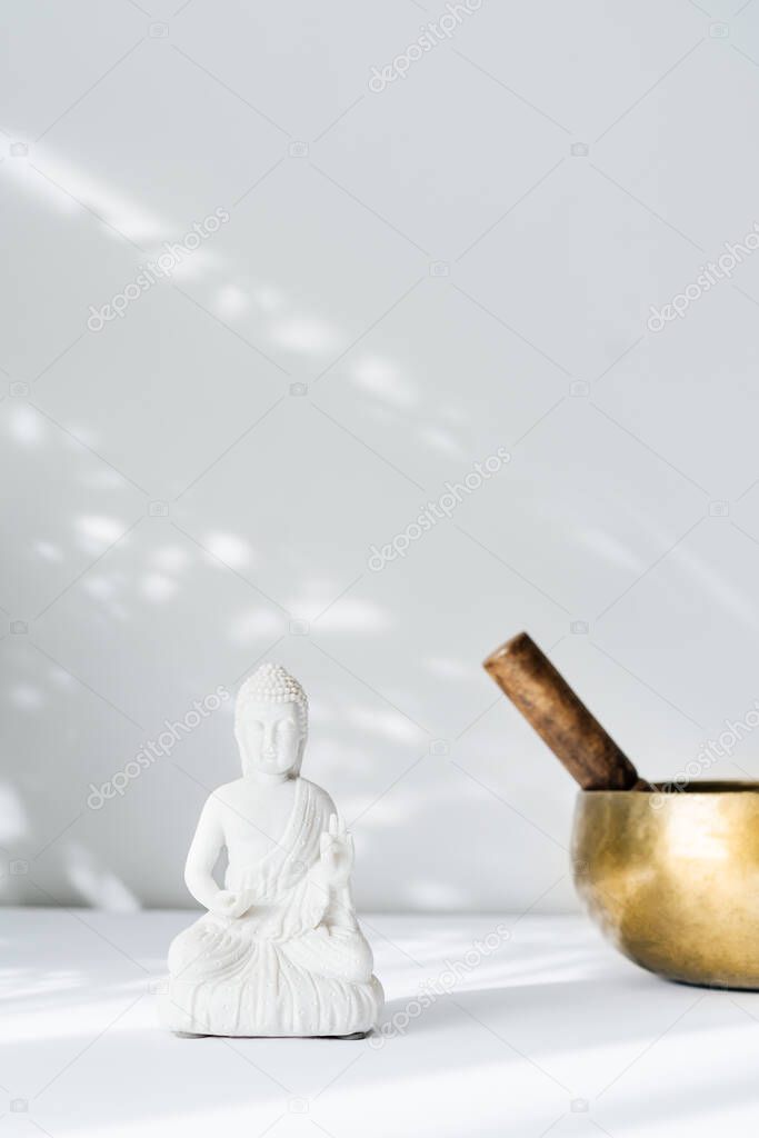 Decorative white Buddha statuette and tibetian singing bowl on the white background with sun light shadows. Meditation and relaxation ritual. Exotic massage. Minimalism. Copy space. Selective focus