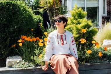 Güneşli bir günde, geleneksel Ukrayna nakışı ve çiçek tarlasının yanında modern pantolon giyen gülümseyen kadın. Modern bir gardıropta etnik kıyafetleri birleştirme kavramı. Moda ve ulusal özgünlük