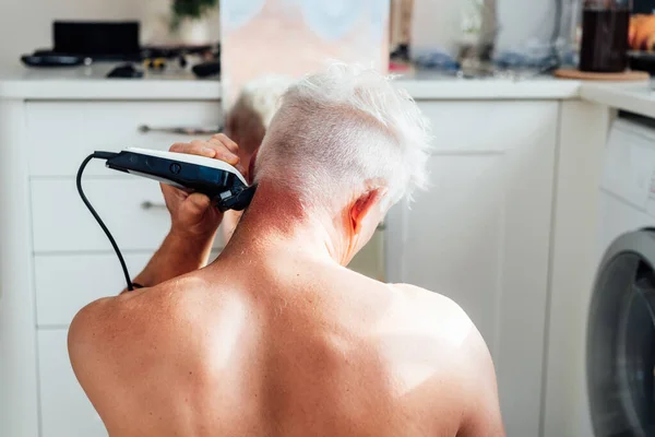 Vista posterior blanco, hombre de pelo plateado haciendo auto corte de pelo con un clipper y se mira en el espejo. autocuidado masculino en casa con afeitadora eléctrica. Enfoque selectivo. — Foto de Stock