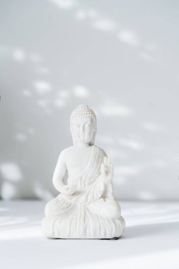Beyaz arka planda, güneş ışığı gölgeli dekoratif beyaz Buda heykelciği. Meditasyon ve rahatlama ayini. Buda 'nın doğum günü. Minimalizm. Uzayı kopyala Dikey kart. Seçici odak.
