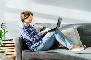 Yumuşak koltukta oturup dizüstü bilgisayar kullanan melez bir kadın gülümsedi. Modern iç mekanda yeşil bitkilerle dolu rahat bir çalışma alanı. Uzaktan kumandalı iş. Çevrimiçi eğitim ya da eğlence. Boşluğu kopyala