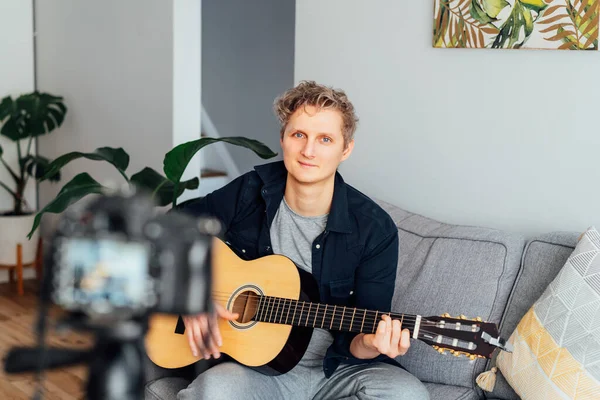 Genç adam akustik gitar çalıyor ve sosyal ağları için müzik videosu kaydediyor. Amatuer müzisyeni modern kamerayla evde içerik yaratıyor. Müzisyen blogcu şarkı söylüyor. hobi etkinlik konsepti.