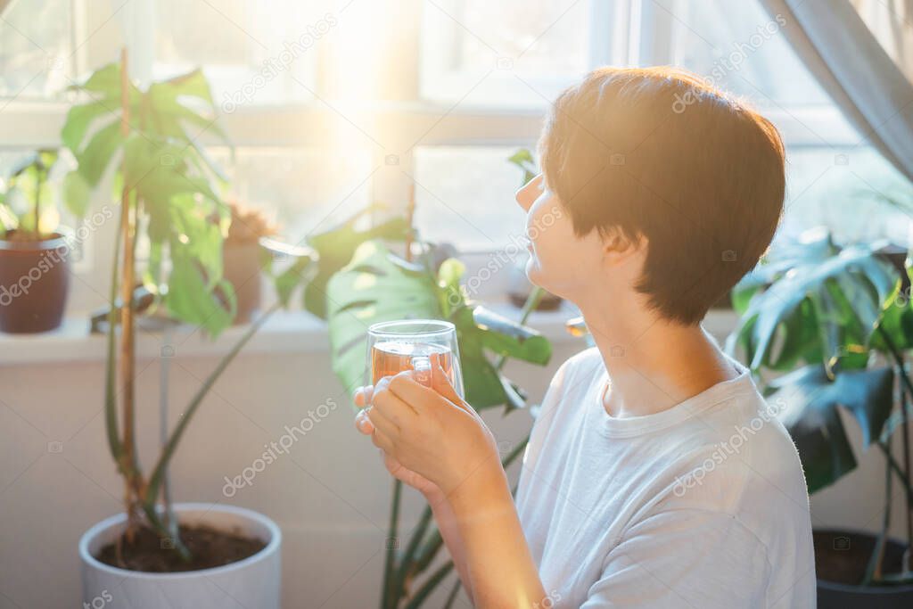 Woman with closed eyes drinking tea in a room with green house plants, enjoying its taste and aroma in a sunset or sunrise light. Relaxing and self-care, personal fulfillment. Selective focus
