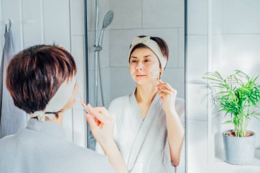 Bornozlu ve saç bantlı bir kadın aynaya bakıyor ve banyoda yeşim taşıyla yüz masajı yapıyor. Cilt bakımı için doğal prosedürler kavramı. Ev Güzeli Kendine Bakım.