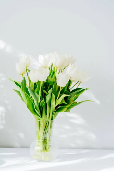 꽃병 안에 하얀 튤립 꽃들 이 꽃다발처럼 피어 있는 봄 부케가 강조와 그림자가 밝은 회색 벽 가까이에 있는 흰 탁자 위에 세워져 있다. 축일 선물, 생일, 3 월 8 일, 어머니 날. 수직 카드입니다. 선택적 초점 — 스톡 사진