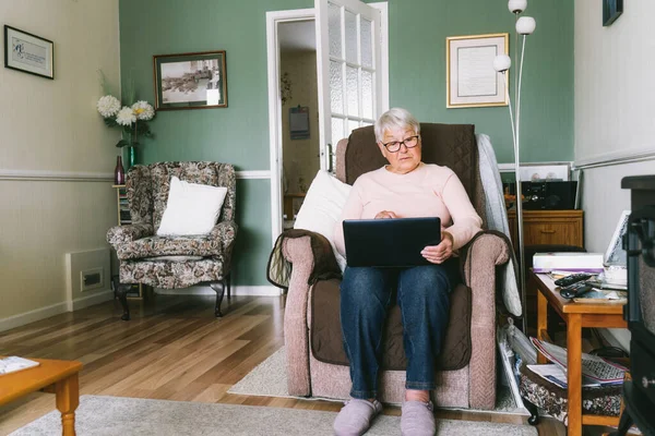 Abuela mayor sentada en un sillón, mirando la pantalla del portátil. Mujer madura de pelo gris leyendo noticias, charlando con la familia, viendo vídeo, consultando en línea con el médico. Mayores y tecnología —  Fotos de Stock