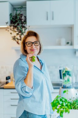 Sağlıklı detoks vegan diyeti. Mutfakta meyve püresi yaparken yeşil kereviz yiyen orta yaşlı kadın portresi. Sağlıklı beslenme, kilo verme programı. Dikey kart. Seçici odak.