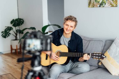 Genç adam akustik gitar çalıyor ve sosyal ağları için müzik videosu kaydediyor. Amatuer müzisyeni modern kamerayla evde içerik yaratıyor. Müzisyen blogcu şarkı söylüyor. hobi etkinlik konsepti