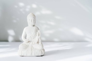 Beyaz arka planda, güneş ışığı gölgeli dekoratif beyaz Buda heykelciği. Meditasyon ve rahatlama ayini. Buda 'nın doğum günü. Minimalizm. Uzayı kopyala Seçici odak