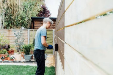 Genç bir işçi bahçede ahşap bir çitle silindirle resim yapıyor. DIY, kendin yap. Ev tadilatı. Ev tadilatı ve yenilenme. Seçici odak, boşluğu kopyala