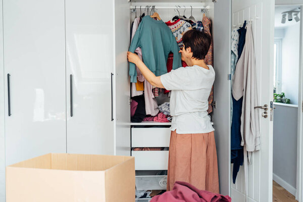 Woman selecting clothes from her wardrobe for donating to a Charity shop. Decluttering, Sorting seasonal clothes and Cleaning Up. Reuse, second-hand concept. Conscious consumer, sustainable lifestyle.