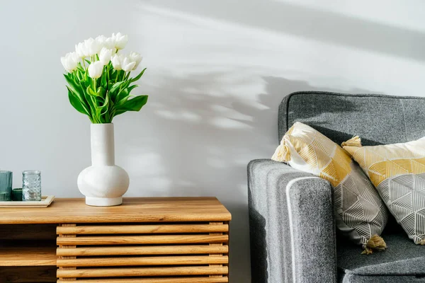 Scandinavian home interior with spring bouquet of white tulip flowers in glass vase, tray with candles standing on a wooden cabinet. Minimalist design with gray sofa and white wall. Springtime.