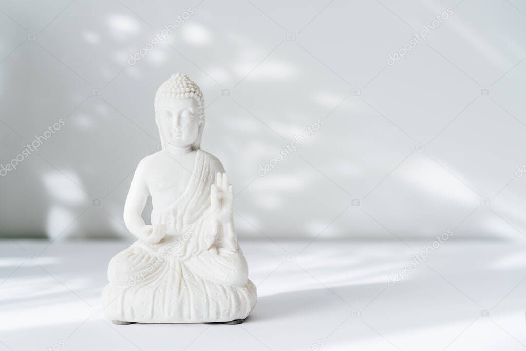 Decorative white Buddha statuette on the white background with sun light shadows. Meditation and relaxation ritual. Buddha birthday. Minimalism. Copy space. Selective focus