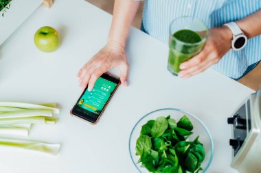 Close up woman with green smoothie fills, making notes in the meal treaker on her phone on the kitchen table. Healthy diet and balanced eating, weight loss online app program. Selective focus.