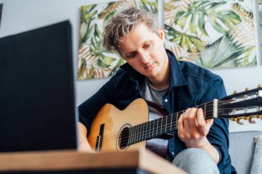 Man taking online guitar lessons with a laptop at home sitting on the gray couch, sofa in the living room. He watching acoustic guitar tutorial on his pc. New hobby. Selective focus.