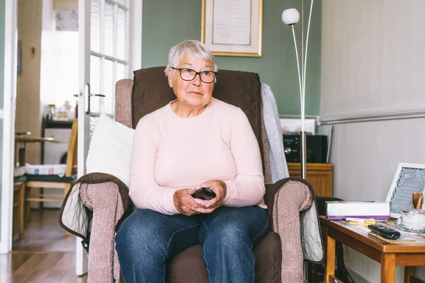 Portrait of retired elderly woman in comfortable armchair with TV remote control and watching television channel in living room. Retirement concept. Selective focus