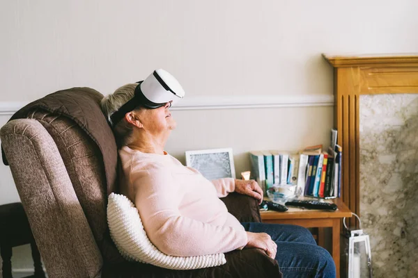Senior woman wearing virtual reality goggles at home. Emotional lady with virtual headset or 3d glasses playing video game. Technology, augmented reality, entertainment industry, metaverse concept