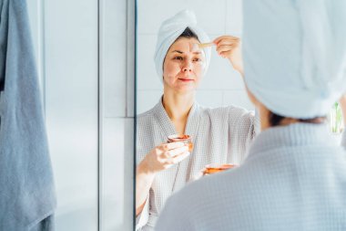 Kafasında havlu olan bir kadın aynaya bakıyor ve banyoda yüzüne kozmetik kil maske takıyor. Cilt bakımı için kozmetik prosedürleri kavramı. Ev Güzeli kişisel bakım