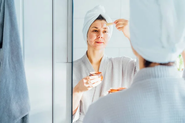 목욕하는 여자는 수건을 머리에 거울을 들여다보며 욕실에서 얼굴에 화장품용 마스크를 — 스톡 사진