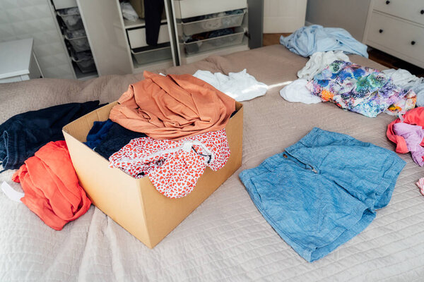 A cardboard box of selected clothes for donating to a Charity shop standing on the bed. Decluttering, Sorting clothes, and Cleaning Up. Reuse, second-hand. Conscious consumer, sustainability