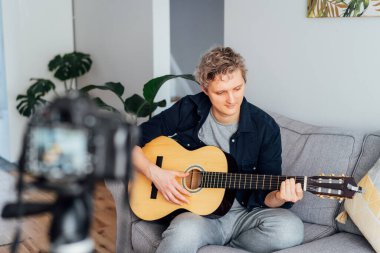 Genç adam akustik gitar çalıyor ve sosyal ağları için müzik videosu kaydediyor. Amatuer müzisyeni modern kamerayla evde içerik yaratıyor. Müzisyen blogcu şarkı söylüyor. hobi etkinlik konsepti.