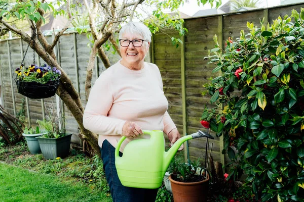 Portrait of happy, smiling senior, mature woman watering flowers with watering can in the garden in a sunny day. Retired elderly woman and gardening hobby. Active Retirement lifestyle
