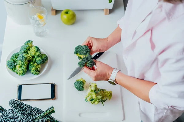 Top view woman watching on her phone with blank white screen while cooking broccoli dish on a kitchen. Online recipes. Mobile app with diet program. Healthy lifestyle, weight loss concept