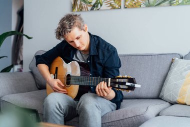 Man taking online guitar lessons via a mobile application on his phone at home sitting on the gray couch, sofa in the living room. He watching acoustic guitar tutorial. New hobby. Selective focus