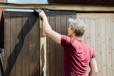 Genç bir işçi fırçayla ahşap bir çit kapısı boyuyor. DIY, kendin yap. Ev tadilatı. Ev tadilatı ve yenilenme. Seçici odak, boşluğu kopyala