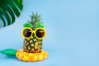 Mavi arka plandaki sarı şişirilebilir yüzüğün üzerinde ananas şeklindeki güneş gözlüğündeki komik ananas. Plaj atmosferinde tropikal bir yaz. Egzotik meyveler. Tatil tatili yaratıcı konsepti. Boşluğu kopyala