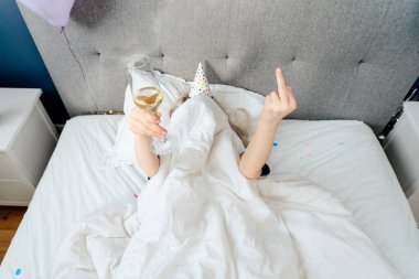 Covered by blanket woman lying on bed with arms raised, holding glass of champagne and showing middle finger, fuck off sign. Passive Birthday plans. Time for yourself. Holiday at home. Happy birthday