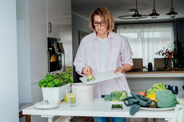 Compost the kitchen waste, recycling. Household woman scraping, throwing vegetables cutting leftovers into the garbage, compost bin while cooking on her kitchen. Environmentally responsible, ecology.