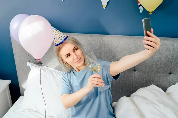 Happy emotional woman in pajama and party cap doing selfie shot on mobile phone at birthday morning while sitting on the bed with champagne and balloons in bedroom. Happy birthday concept.