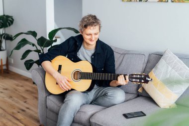 Adamın biri telefonuyla internetten gitar dersi alıyor. Gri koltukta oturuyor. Oturma odasındaki kanepe. Akustik gitar dersi izliyor. Yeni hobi. Seçici odak