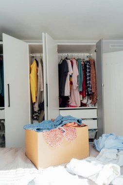 Yatağın üzerinde duran bir hayır kurumuna bağışlamak için seçilmiş kıyafetlerin olduğu bir karton kutu. Temizlik, giysi ayıklama ve temizlik. Tekrar kullan, ikinci el. Bilinçli tüketici, sürdürülebilirlik