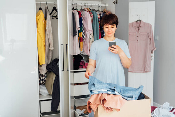 Женщина фотографирует шорты, используя свой смартфон для продажи или пожертвования своей одежды. Раздевание, сортировка одежды и уборка гардероба. Повторное использование, подержанная концепция. Сознательный потребитель