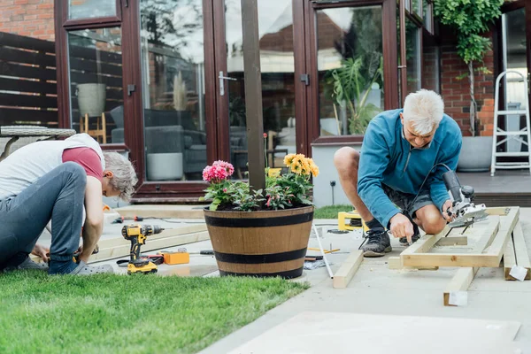 Adult son and father carpenter working together, handle wooden timbers in the garden. Patio construction by your own. DIY, Do it yourself. Home renovation, improvement, refurbishment. Selective focus.