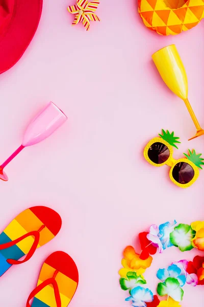 Flat lay of summer vibes concept with colorful pool party items, funny sunglasses, cocktail glasses. inflatable drink holders, flip flops, bucket hat, flower necklaces on pink background. Copy space