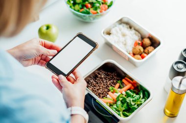 Healthy diet plan for weight loss, daily ready meal menu. Woman using phone with blank screen for app while weighing lunch box cooked in advance on kitchen scale. Balanced portion. Pre-cooking.