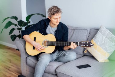 Adamın biri telefonuyla internetten gitar dersi alıyor. Gri koltukta oturuyor. Oturma odasındaki kanepe. Akustik gitar dersi izliyor. Yeni hobi. Seçici odak