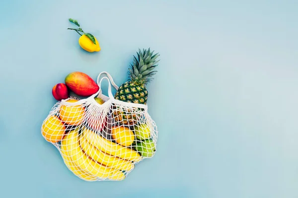 Top view string bag, mesh bag, grocery bag with organic fresh exotic fruits spilling from a reusable shopping bag on light blue background. Zero waste, eco-friendly shopping. Healthy diet. Flat Lay.
