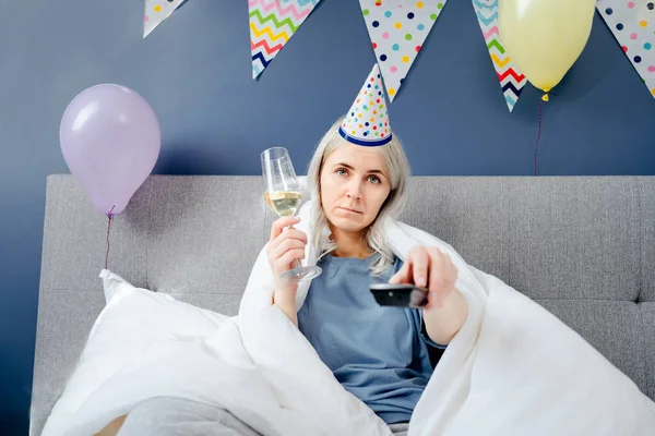 Sad woman in pajamas, party cap drinks champagne and watches TV, wrapped in a blanket while sitting on a bed in a decorated bedroom. Celebrates a birthday alone. Boring home party. Selective focus.