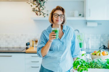 Smiling middle aged woman with just made glass of detox shake, green smoothie in the kitchen. Healthy dieting, eating, cooking. Natural anti aging methods, weight loss program. Vegan, vegetarian diet clipart