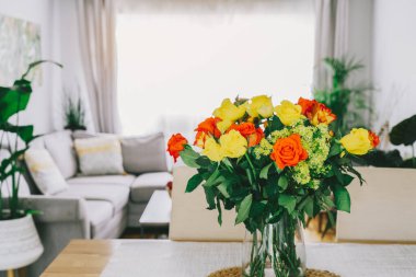 Kitchen counter table with focus on vase with yellow and orange roses on it with blurred background of modern cozy living room with couch and green plants. Open space home interior design. Copy space